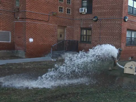 Water pumped out of a flooded basement gushing across the grass beside red brick apartment buildings