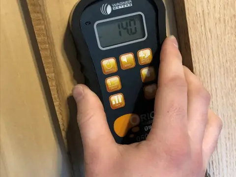 A hand pressing a pin moisture meter against a board to read how wet it is