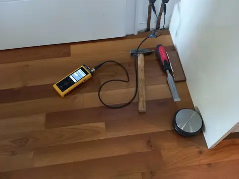 A moisture meter on a wood floor, its probe cable running to a white wall