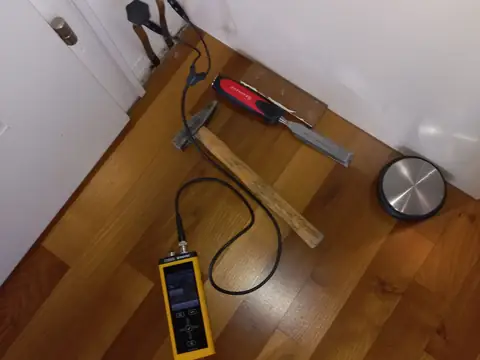A moisture meter and its probe set on a wood floor at the base of a wall