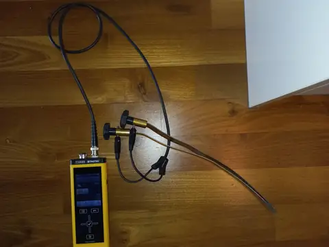 A moisture meter on a wood floor beside a wall, its cable running to a probe