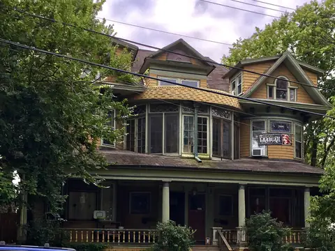 A large Victorian house with a deep front porch behind trees in Westerleigh, Staten Island
