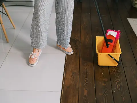 A mop and a yellow bucket of water on a dark wooden floor beside a tiled one